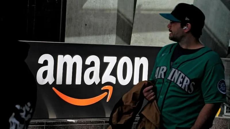 A photograph capturing a man wearing a green Seattle Mariners jersey standing beside a large, backlit Amazon corporate sign. The image represents the company's presence in Seattle, reflecting its corporate identity during a period of business news regarding layoffs.
