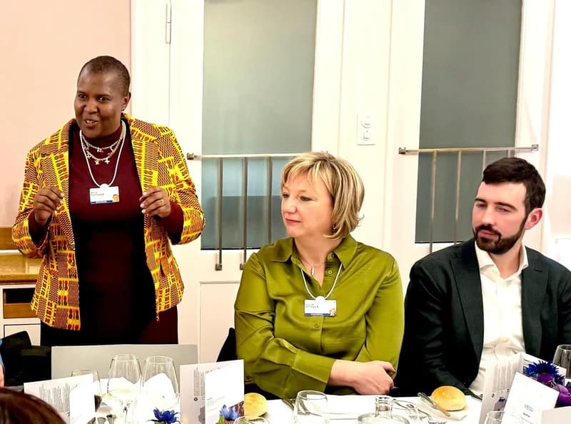 Dr. Agnes Kalibata, President of AGRA, is captured standing and speaking at a formal dinner or meeting event. She wears a vibrant yellow and maroon patterned jacket over a maroon dress, while two seated colleagues, a woman in green and a man in a suit, listen attentively.
