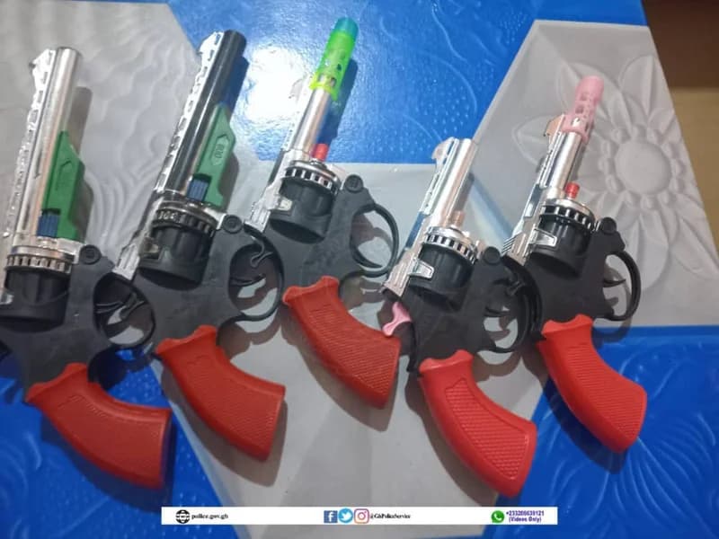 The image shows five plastic toy revolvers with distinctive red handles and silver-colored barrels laid out on a blue and white patterned surface. A banner at the bottom contains the official website and social media handles for the Ghana Police Service, suggesting a public awareness campaign.