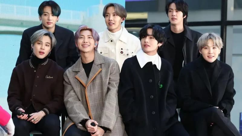 The image features all seven members of the South Korean boy band BTS posing together. They are dressed in stylish autumn or winter clothing, including wool coats and turtlenecks, with Jimin holding a microphone while seated.