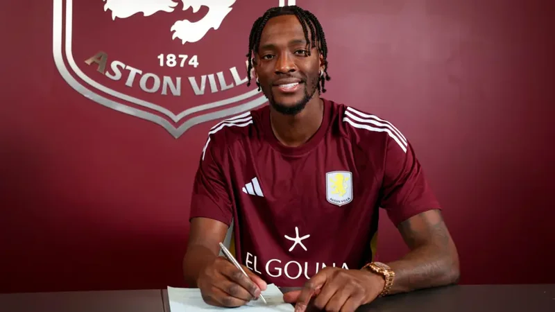 Footballer Tammy Abraham is pictured smiling while signing a contract at Aston Villa. He is dressed in the club's claret training kit and stands in front of a wall featuring the official Aston Villa crest and the year 1874.