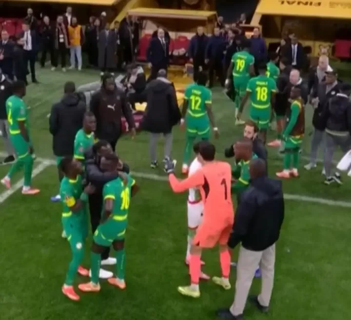The image depicts a crowded football pitch following a match, showing players in green kits and a goalkeeper in orange amid a scene of activity. Various officials and staff members are visible in the background near the team dugouts, suggesting a post-game gathering or incident.