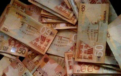 A close-up photograph showing a scattered pile of Ghanaian 50 Cedi banknotes. The currency features the 'GHANA' text and the '50' denomination clearly visible on several notes against a dark background.