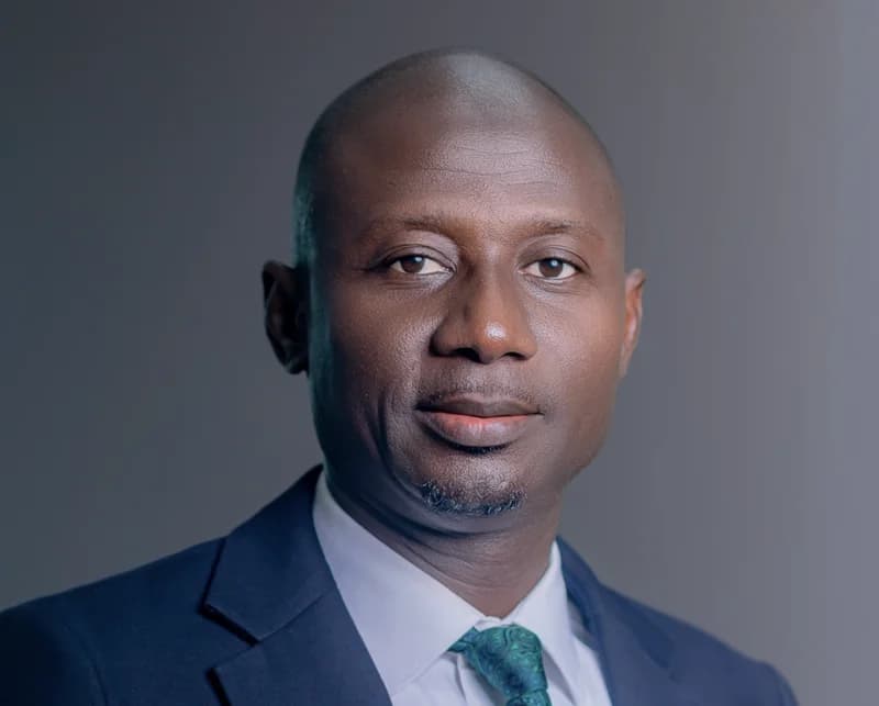 A professional portrait of a man of African descent wearing a dark navy suit, a white dress shirt, and a patterned green necktie. He is positioned against a solid grey background, looking directly into the camera with a professional and composed expression.