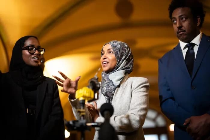 Congresswoman Ilhan Omar is seen speaking at a microphone during a press conference or public event, gesturing with her right hand. She is flanked by two people, a woman in a black hijab and glasses on her left, and a man in a blue suit on her right, in a warmly lit indoor setting.