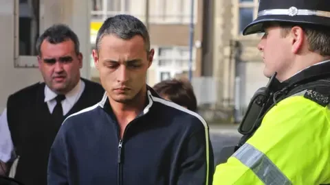 PA Media Paul Grabham arriving at Swansea Magistrates Court, where he is charged with the murder of his wife, Kirsty. Paul is wearing a dark blue zip up top with white stripes down the shoulders and sleeves and is staring at the ground as he walks. A prison guard is walking behind him wearing a white shirt with a black tie and a gilet style jumper over the top. To Paul's immediate left is a police officer in full uniform looking at him