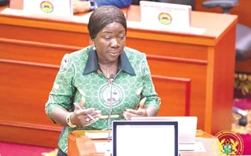 A woman wearing a green dress with the Ghana Revenue Authority logo speaks at a podium in a formal setting, possibly the Parliament of Ghana. She is gesturing while addressing an audience, with wooden furniture and official emblems visible in the background.