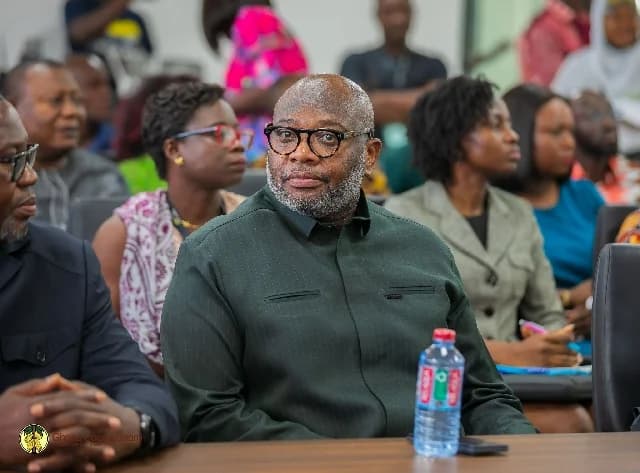 The image shows Joseph Boahen Aidoo, the Chief Executive of the Ghana Cocoa Board (COCOBOD), seated at a table during what appears to be a press conference or official meeting. He is wearing glasses and a dark green shirt, with a bottle of water on the table in front of him and several other participants visible in the background.