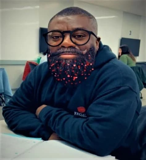 A photograph showing Professor Ransford Gyampo, a prominent Ghanaian political science lecturer, wearing black glasses and a dark hoodie with 'REGAL' text. He is seated at a desk, and his beard has been digitally edited to include small red heart-shaped patterns.