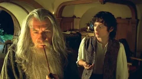 New Line via Getty Images Ian McKellen on the left is wearing a grey hooded robe holding a smoking pipe to his mouth. He has long grey straight hair and a long grey beard. Elijah on the right is looking at Ian. He has brown curly hair and is wearing a white shirt and open brown waistcoat.