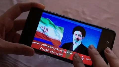 AFP via Getty Images Hands holding a mobile phone showing an image of Iran's new supreme leader Mojtaba Khamenei on 12 March 2026, as statement by him is broadcast.