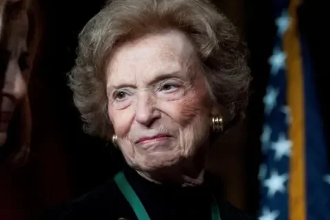 Reuters Doris Fisher pictured in 2011, wearing gold earrings, a black top, against a dark background with the American flag seen behind her