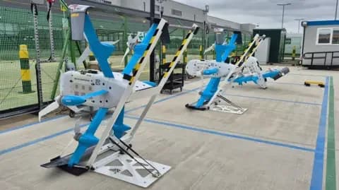 Two Amazon drones - grey and blue unmanned aircraft - in a concrete compound with blue and green boxes painted on the ground