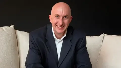 Tim Beasley Tim Beasley smiles at the camera. He is wearing a dark suit and set on a white sofa
