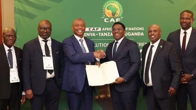 A group of four sports ministers and football bosses from Tanzania and Uganda, all wearing dark suits, white shirts and dark ties, pose for a photo alongside Confederation of African Football president Patrice Motsepe (third from left) and secretary general Samson Adamu (far right), who are similarly dressed. The group stands in front of a green board which features a Caf logo and Motsepe is shaking hands with one of the administrators as the pair hold open a large document for the camera