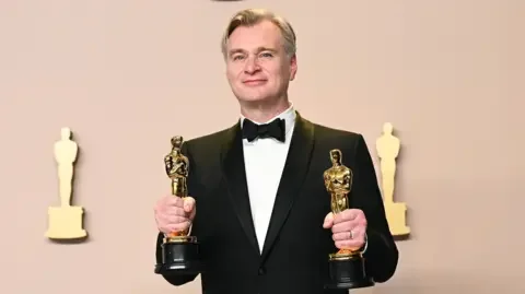 Getty Images Christopher Nolan smiling in a black bow tie and dinner jacket, holding an Oscar statuette in each hand, in front of an Oscars backdrop