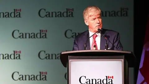 Getty Images An image of Tim Hodgson, Canada's minister of natural resources, speaking at a podium. Behind him is a backdrop with a repeated Canada logo, featuring the word Canada and a small Canadian flag above the letter 'a'. He is wearing a suit, and has combed over grey hair.