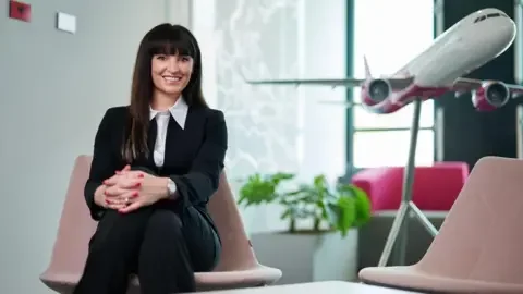 Wizz Air Yvonne smiling while wearing a suit and sitting in a pale pink chair, in front of a large model of an airplane.