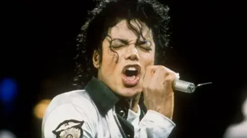 US singer Michael Jackson performing at a 1998 concert in London. He is wearing a white navy jacket with a white collar and singing into a microphone with his eyes closed
