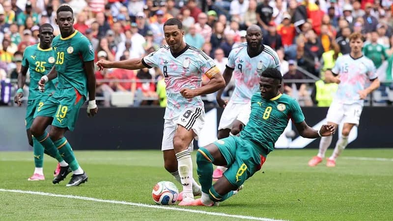 Belgium captain Youri Tielemans is fouled by Lamine Camara during the 2026 World Cup last-32 tie in Seattle. Tielemans is standing with his left leg planted forward as Camara slides in from the side, leading with his right leg. The contact between the pair is difficult to see, with the ball positioned right in front of their pink boots. Behind them, watching on, are two Senegal defenders and two Belgium attackers, including Romelu Lukaku. The Belgians wear white socks and shorts and light shirts made up of pink and turquoise patches, while the Senegalese are all in green with red and yellow trim. A blurred section of crowd is visible in the background