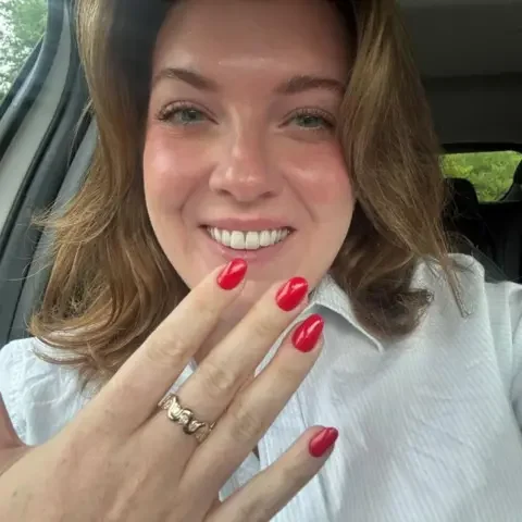 Alex Proie Alex Proie sitting in a car holding up her hand with a gold and diamond wave design ring on her middle finger and smiling