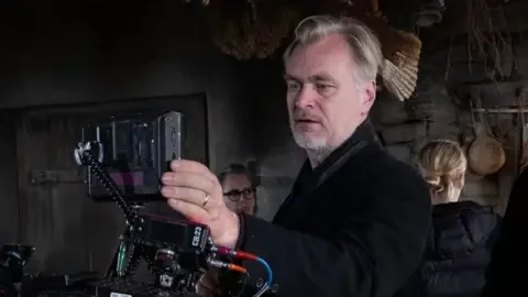 Universal Pictures Christopher Nolan in the centre of the photo and is looking through an Imax camera. He is wearing a black jacket.