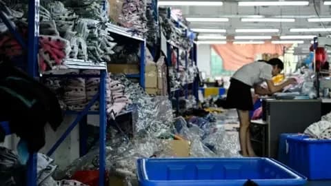 AFP via Getty Images A worker makes clothes at a garment factory that supplies Shein. She is surrounded by piles of clothes stacked on blue shelves.