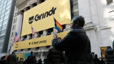 Getty Images A woman talks to someone on a video call on her smartphone in front of the building housing the New York Stock Exchange on Wall Street, which is covered in a huge Grindr poster to mark the company going public in November 2022. A LGBTQ+ flag is flying beneath it, alongside the US flag, with people milling around below.