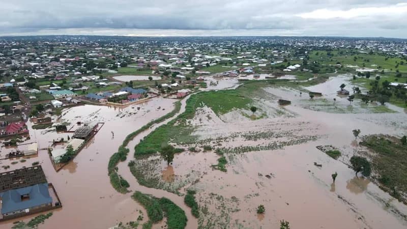An aerial view shows a residential community and surrounding farmlands heavily inundated by muddy brown floodwaters. The image captures the scale of the natural disaster, with water reaching the walls of houses and submerging large tracts of land under a gloomy, overcast sky.