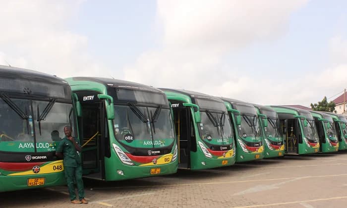A row of green Aayalolo rapid transit buses is parked at a terminal or bus depot in Accra. A staff member wearing a green uniform is leaning against the front of the lead Scania bus, which clearly shows the fleet number 048 and registration plate GE 9134-16.