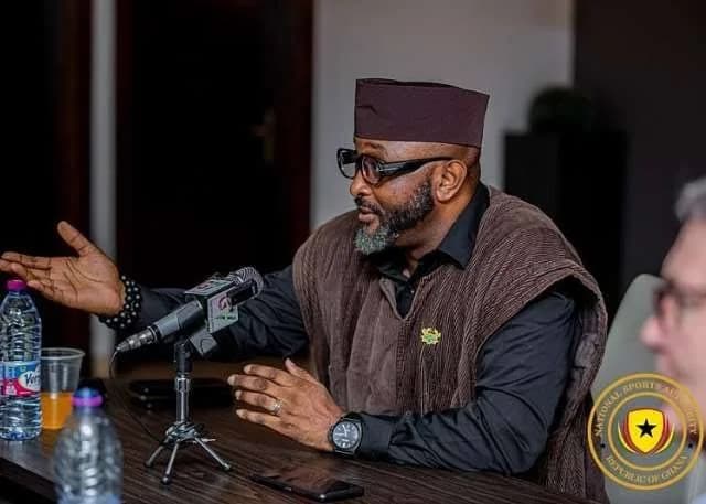 A man, identified as a senior official of Ghana's National Sports Authority (NSA), is seen speaking at a press engagement. He is seated at a table with microphones and water bottles, wearing a distinctive brown cap and traditional smock, with the NSA logo visible in the foreground.