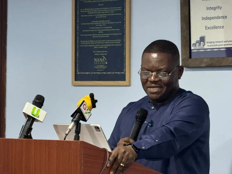 Gayheart Mensah is seen speaking at a podium during an event, likely related to journalism or governance. He stands behind several microphones, including those from UTV and GTV, with a CDD-Ghana poster emphasizing 'Integrity, Independence, Excellence' visible in the background.