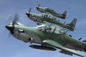 Three military turboprop aircraft, appearing to be Embraer EMB 314 Super Tucanos, fly in a tight, staggered formation against a clear blue sky. The planes are painted in a green and grey camouflage pattern with visible pilots inside the cockpits and external fuel tanks mounted under the wings.