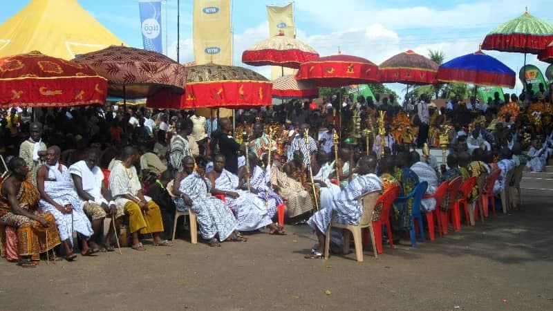A large gathering of traditional leaders and community members seated under vibrant state umbrellas during the Odwira festival celebration. The participants are dressed in traditional Ghanaian patterned wraps and cloths, while corporate banners from MTN and Tigo are visible in the background.