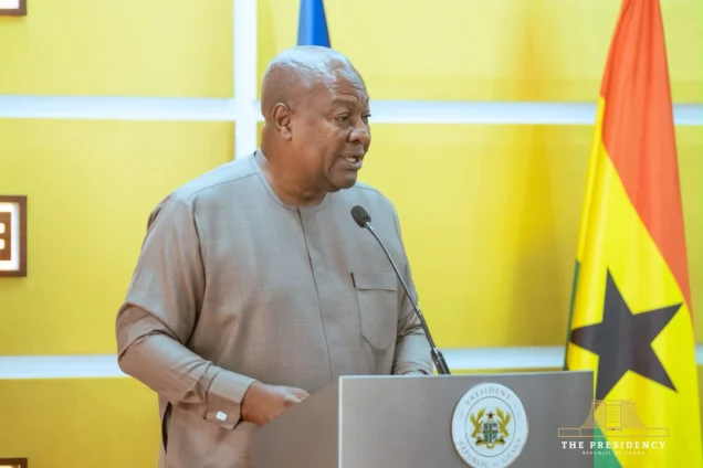 President John Dramani Mahama is captured speaking at an official event, standing behind a podium marked with the Presidential Seal of Ghana. The background features a vibrant yellow wall and the national flag of Ghana, indicating a high-level government address.