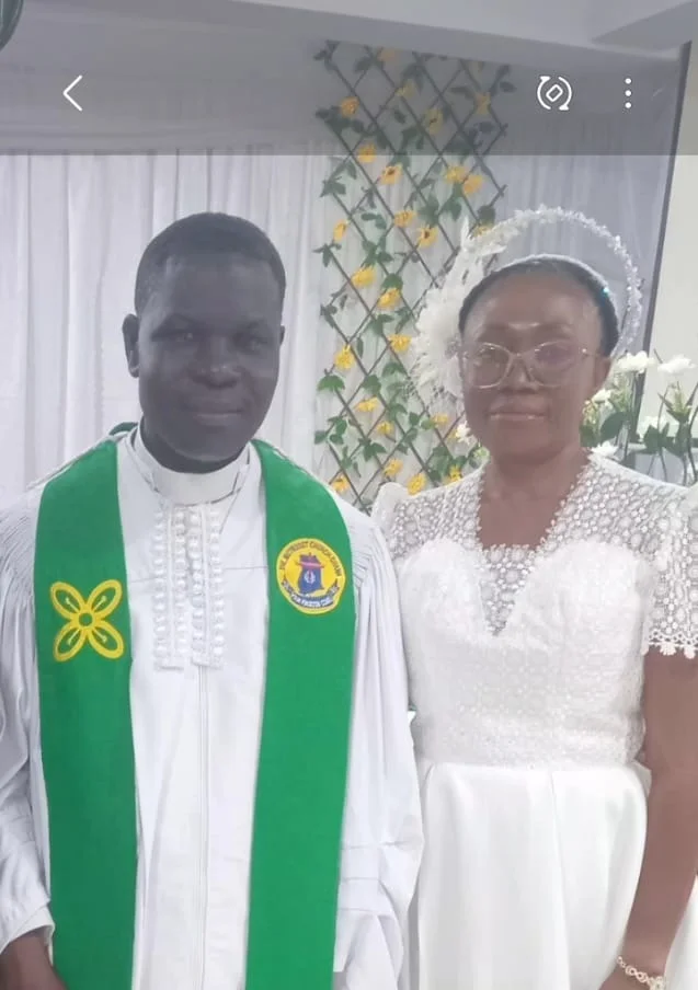 A photograph of Reverend Seth Williams in Methodist clerical robes standing alongside a woman in formal white attire. The pair is posed in front of a white curtain and floral trellis background, likely during a church ceremony or celebration of his service.