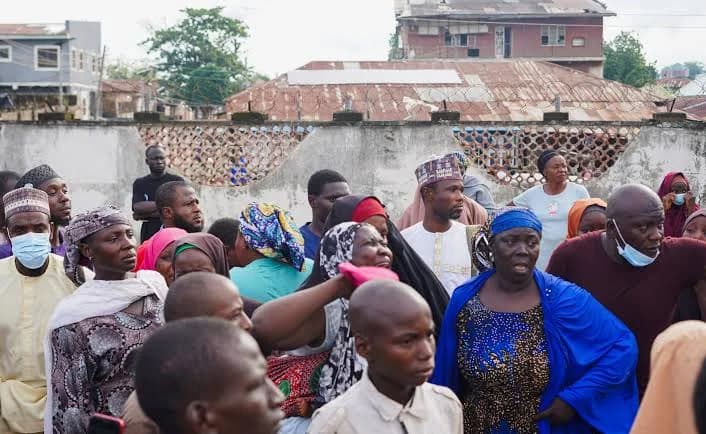 A large group of mourning and distressed individuals gather outdoors in what appears to be a residential area in a Nigerian city. Many participants are wearing traditional attire, including hijabs and caps, while several individuals are seen wearing blue surgical face masks amidst the somber crowd.