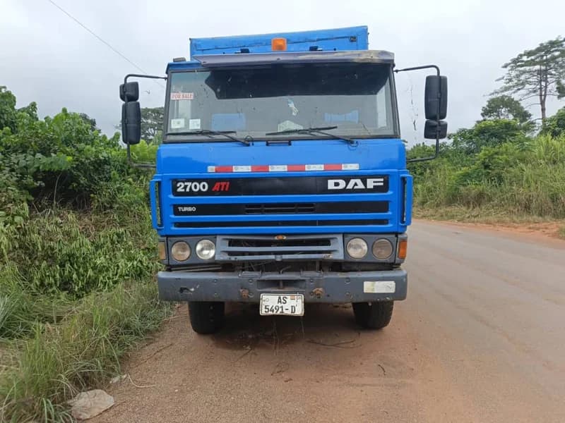 A blue DAF 2700 ATi truck is parked on the side of a paved road, partially obstructing the shoulder. The vehicle features a Ghanaian license plate and a 'FOR SALE' sign on the windshield, set against a backdrop of dense tropical greenery. This image illustrates the abandoned truck that residents are concerned about on the Benchemaa-Oseikojokrom highway.