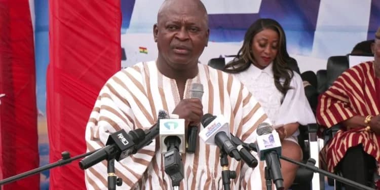 A man wearing a traditional brown and white striped Ghanaian smock (batakari) speaks into multiple microphones at a press conference. He is flanked by other attendees in the background against a backdrop that includes a small Ghana flag and a red curtain.