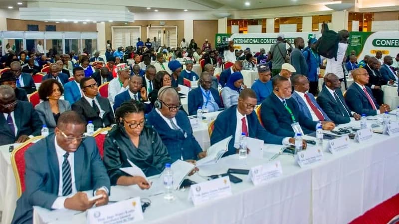 A wide-angle photograph of a large international conference hall filled with delegates and officials. Participants are seated at long, white-clothed tables, many wearing formal business attire or traditional West African garments while listening to proceedings.