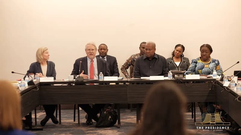 President John Mahama sits on a panel with other dignitaries during a high-level meeting, likely associated with the UN General Assembly. He is seen listening attentively while a colleague in a suit and red tie speaks into a microphone at a long conference table.