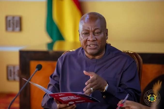 President John Dramani Mahama is pictured during a formal meeting, holding a document and gesturing with his hand as he speaks. He is seated behind a desk with a microphone, with the Flag of Ghana and the presidential seal visible in the background.
