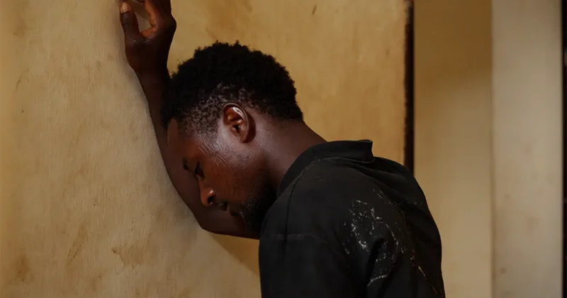 A close-up photograph of a young Black man leaning his head and arm against a textured, light-colored wall in a moment of visible distress or exhaustion. He is wearing a dark shirt, and the low lighting emphasizes the somber and contemplative mood of the scene.