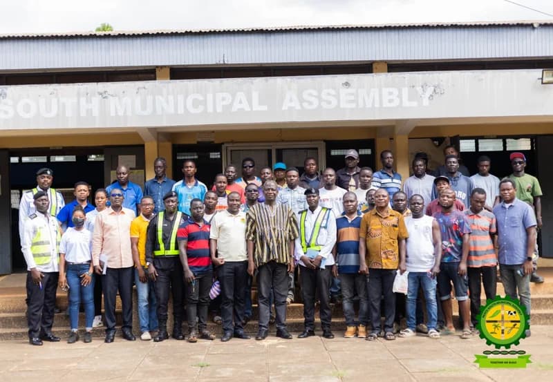 A large group of people, including Ghana Police Service officers and local residents, pose for a photograph in front of the Akatsi South Municipal Assembly building. The gathering represents a road safety sensitization event aimed at educating road users on new regulations in the municipality.