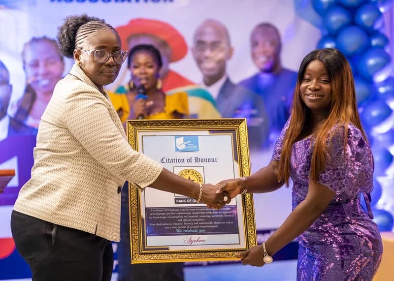 A woman in a beige blazer and glasses presents a framed 'Citation of Honour' to another woman in a sparkly purple dress. The award is from the Bono Ghana Journalists Association (GJA) and is being presented to the Bank of Ghana in recognition of their support for media development and financial literacy workshops.