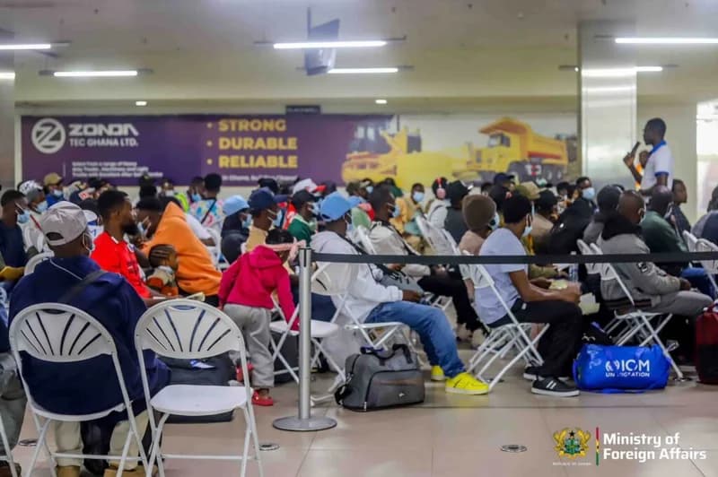 A large group of repatriated Ghanaians, many wearing face masks and hats, are seated on white folding chairs in what appears to be an airport arrival hall or processing center. An International Organization for Migration (IOM) bag is visible on the floor, while a person addresses the group from the side.