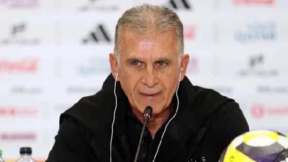 Football manager Carlos Queiroz is pictured during a press conference, wearing earpieces and speaking into a microphone. The background consists of a branded media wall featuring various sports and corporate sponsors.