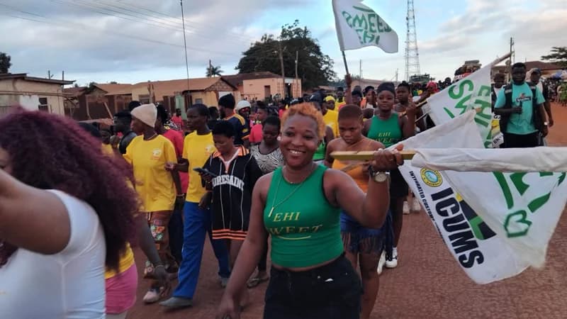 A vibrant crowd of residents in Ehi participate in a street procession to promote the 10th Nutome Gali Za celebration. Many participants, mostly youth, are seen wearing green and yellow t-shirts and carrying flags with branding such as 'Ehi Sweet' and 'ICUMS' while walking through a residential area.