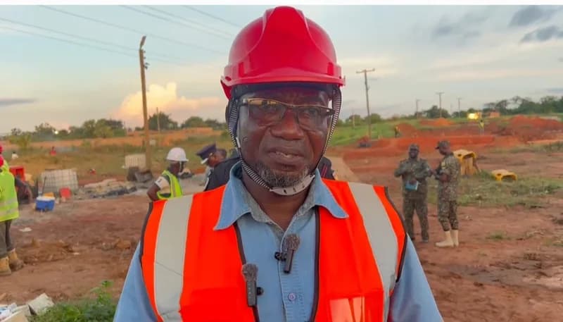 A man wearing a red hard hat and an orange safety vest stands at an outdoor construction site, likely providing an update on a development project. In the background, several individuals including security personnel in camouflage and workers are visible on a site with red earth and utility poles.