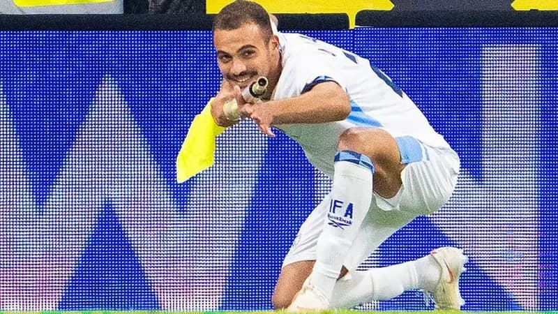 A football player in a white Israel national team kit kneels on the pitch while holding a yellow corner flag. He is mimicking a shooting motion with his hands and the flag pole as part of a goal celebration against a backdrop of a blue digital advertising board.